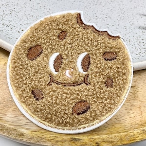 Op de afbeelding: Een bruine, pluche koekje met chocolade chips en een glimlachend gezicht. Het koekje heeft een hap eruit.