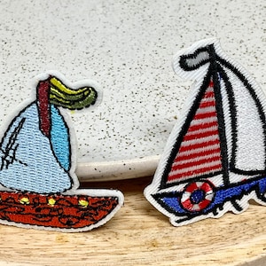Peut inclure: Deux patchs brodés thermocollants représentant des voiliers. Un voilier est bleu et blanc avec une voile rouge et blanche à rayures. L'autre voilier est rouge, blanc et bleu avec une voile jaune.