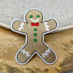 Peut inclure: Un patch brodé en forme d'homme en pain d'épices avec un nœud papillon rouge et des boutons verts. Le patch est beige avec des détails de glaçage blanc.