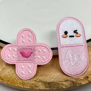 May include: Two embroidered patches: a pink bandage with a heart and a pink and white pill with a smiling face and a heartbeat line. The patches are on a wooden tray with a white plate.
