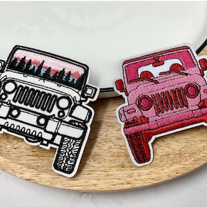 May include: Two embroidered patches of a Jeep. One is outlined in black and white with a mountain scene across the top. The other is pink and red. Both patches are on a wooden plate.