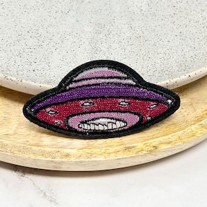 Peut inclure: Un patch brodé noir, violet et rouge représentant un OVNI avec un visage souriant.