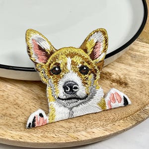 Puede incluir: Parche bordado de un perro marrón y blanco con orejas rosas que asoma por detrás de una superficie de madera.