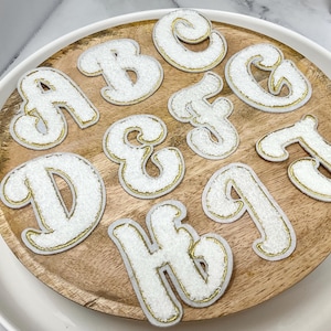 May include: A collection of white chenille letter patches featuring a gold glitter border. The set includes the letters A, B, C, D, E, F, G, H, I, and J, each rendered in a bold, rounded font style with a textured surface and metallic trim.