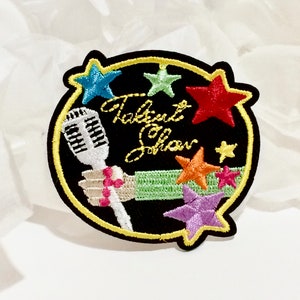 May include: A black and yellow embroidered patch featuring a microphone, a hand holding the microphone, and the text "Talent Show" surrounded by colourful stars.