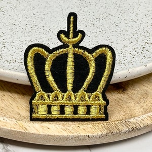 Peut inclure: Un écusson brodé d'une couronne dorée sur un fond en feutre noir. La couronne a un sommet pointu et cinq pointes.