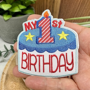 May include: A white embroidered patch featuring a blue and white cake with a red and white striped candle with a flame on top. The cake has two yellow stars on it. The text "MY 1ST BIRTHDAY" is embroidered in red on the patch.