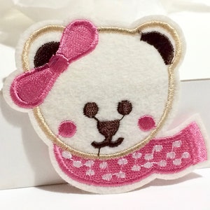 Op de afbeelding: Witte vilt applique van een beer met een roze strik en een roze-witte stippen sjaal.