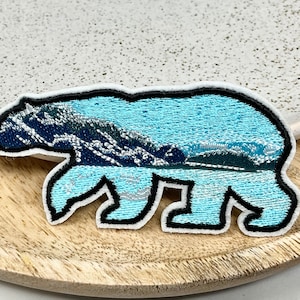 Può includere: Toppa ricamata a forma di orso, con una scena di montagna e lago. L'orso è azzurro con un contorno nero e un bordo bianco. La scena della montagna presenta sfumature di blu, verde e bianco.