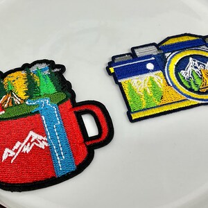 Peut inclure: Deux écussons brodés : une tasse rouge avec une scène de montagne et un écusson d'appareil photo bleu et jaune. L'écusson de la tasse représente une cascade et une chaîne de montagnes. L'écusson de l'appareil photo présente un motif de montagne et d'arbres.