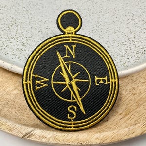 May include: Black and gold embroidered compass patch. The compass rose features the cardinal directions, with a gold needle pointing north. The patch has a gold border and a loop at the top.