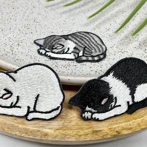 Peut inclure: Trois patchs brodés thermocollants représentant des chats endormis. Les chats sont enroulés sur eux-mêmes, les pattes sur le visage. Un chat est gris et blanc, un autre est noir et blanc et le dernier est blanc.