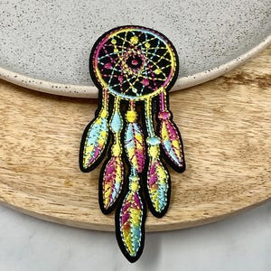 May include: A colourful dreamcatcher with a black background and feathers in shades of blue, pink, and yellow. The dreamcatcher is embroidered with a geometric pattern.