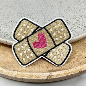 Peut inclure: Un patch brodé marron et blanc en forme de pansement avec un cœur rose au centre.