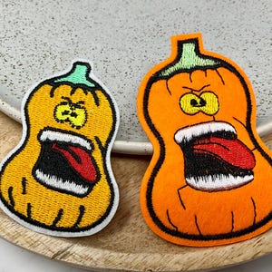 Peut inclure: Deux patchs de citrouille en feutre brodés avec des expressions en colère et la bouche ouverte. Les citrouilles sont orange et jaunes avec des yeux noirs et des dents blanches. Une citrouille a la langue rouge qui sort.
