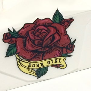 Peut inclure: Illustration détaillée d'une rose rouge avec des feuilles vertes. Une bannière jaune avec les mots "ROSE GIRL" est en dessous de la rose. Le design est sur fond blanc.
