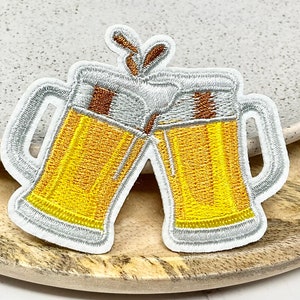 Op de afbeelding: Twee witte geborduurde bierglazen die klinken met geel bier erin en een bruin schuim bovenop. De glazen staan op een wit en bruin oppervlak.