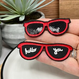 Peut inclure: Deux patchs thermocollants brodés en forme de lunettes de soleil, de couleur rouge et noire. Le patch du haut est orné de paillettes argentées. Le patch du bas porte l'inscription "hello you" en blanc.