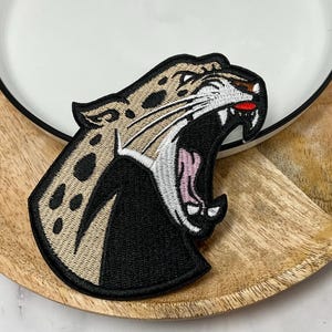 Peut inclure: Un écusson brodé représentant la tête d'un léopard en profil, gueule ouverte. Le motif utilise du fil beige pour le pelage avec des taches noires, du fil blanc pour le museau et les dents, une langue rose et une bordure noire tout autour.
