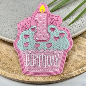 May include: Pink embroidered iron-on patch with a cupcake design. The cupcake has a green frosting with pink hearts and a pink candle with a white flame. The text "My 1st Birthday" is embroidered on the patch.