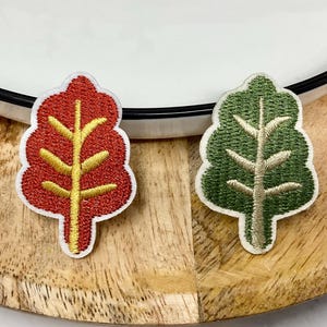 Peut inclure: Deux écussons brodés en forme d'arbre. L'un est rouge avec une tige et des branches jaunes, l'autre est vert avec une tige et des branches beiges. Les deux ont des bordures blanches. Sur une surface en bois, près d'une assiette blanche.