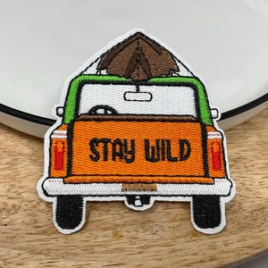 May include: Embroidered patch depicting an orange truck with a brown tent on its roof. The truck has a green roof and the words "STAY WILD" in black. The patch is white-bordered, suitable for application on clothing or accessories.
