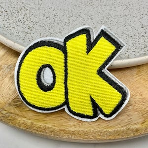 Op de afbeelding: Gele vilt patch met zwarte omtrek in de vorm van het woord "ok" in een cartoon lettertype.