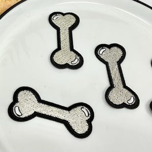 May include: Four white embroidered bone-shaped patches on a white plate. The patches are outlined in black thread.