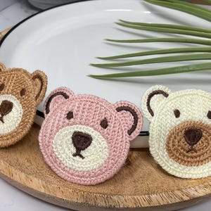 Peut inclure: Trois appliqués en forme de tête d'ours au crochet, dans les tons de marron, de rose et de crème, chacun avec un museau blanc et des détails marron foncé. Les appliqués sont disposés sur un plateau en bois avec une assiette blanche et des feuilles vertes en arrière-plan.