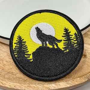 Op de afbeelding: Een ronde geborduurde patch met een zwart silhouet van een huilende wolf op een rotsachtige klif. De achtergrond bestaat uit een heldergele lucht met een witte volle maan en zwarte dennenbomen aan weerszijden, omrand door een zwarte rand.