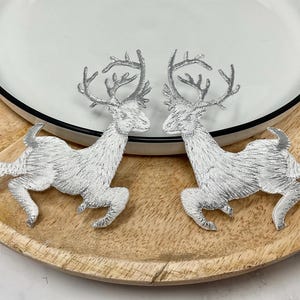 Könnte beinhalten: Zwei silberne, gestickte Rentierfiguren mit detaillierten Geweihen und Beinen werden auf einem Holztablett präsentiert. Die Rentiere sind einander zugewandt und stehen auf einem weißen Teller mit schwarzem Rand. Das Tablett hat eine natürliche Holzmaserung.