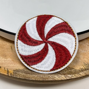 May include: A circular embroidered patch featuring a red and white peppermint candy swirl design. The patch has a white border with gold-coloured stitching around the edge, creating a distinct spiral pattern that radiates from the centre.