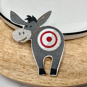 May include: Embroidered iron-on patch of a gray donkey with a red and white target on its rear end.