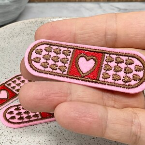 Peut inclure: Patch brodé en feutre rose en forme de pansement avec un cœur rouge au centre. Le pansement est décoré de cœurs brodés marrons.