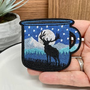 Puede incluir: Parche bordado con forma de taza, que representa una escena nocturna con un ciervo, montañas y una luna llena. El parche tiene un borde negro y es principalmente azul, blanco y negro.