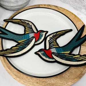 Puede incluir: Dos parches bordados de golondrinas con detalles en dorado, azul, rojo y blanco. Los parches están sobre un plato blanco con borde de madera.
