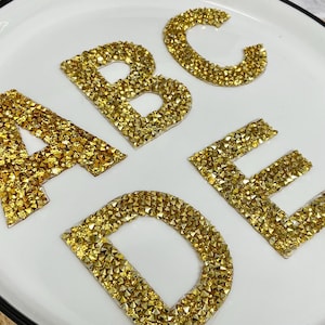 May include: Close-up of the letters A, B, C, D, and E, each encrusted with small, sparkling gold-coloured crystals. The letters are arranged on a white plate with a black rim. The letters are a decorative element.