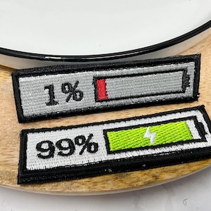 Peut inclure: Deux écussons brodés avec un motif de niveau de batterie. L'un affiche 1% avec une barre rouge, l'autre affiche 99% avec une barre verte et un éclair. Les deux ont des bordures noires et du texte blanc.