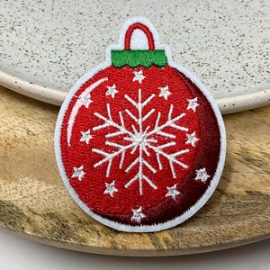 Peut inclure: Boule de Noël rouge avec un flocon de neige blanc et des étoiles blanches. La boule est brodée et possède une boucle en feutre vert.