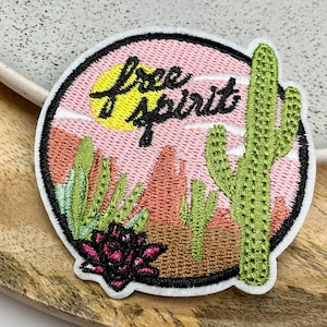 Peut inclure: Patch brodé avec un paysage désertique rose, un soleil jaune et le texte "free spirit". Le patch présente un grand cactus vert et une petite fleur rose.