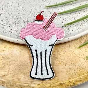 Peut inclure: Un patch brodé représentant un milk-shake rose et blanc avec une cerise rouge sur le dessus et une paille rouge et blanche rayée.