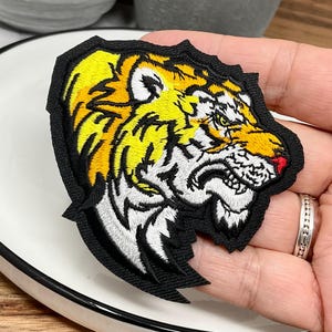 Op de afbeelding: Een geborduurde patch met een grommende tijgerkop in profiel. Het ontwerp gebruikt levendig geel, wit en zwart garen met een klein rood accent op de neus, allemaal omlijnd door een dikke zwarte rand op een donkere stoffen ondergrond.