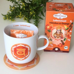 Op de afbeelding: Een witte mok met een oranje chakra-symbool en de tekst "I am creative!" De mok staat op een bijpassende oranje onderzetter. Op de achtergrond staat een doos "SHOTI MAA Lebenslust Hibiskus & Orange" thee.
