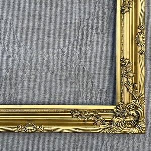 May include: A gold ornate picture frame with intricate carvings and a raised edge. The frame is against a gray textured background.