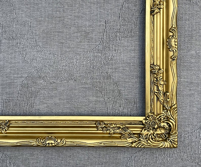 May include: A gold ornate picture frame with intricate carvings and a raised edge. The frame is against a gray textured background.