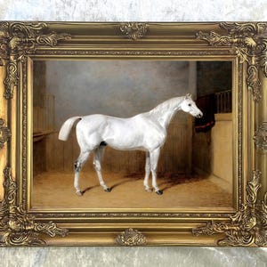May include: A framed oil painting of a white horse standing in a stable. The horse is facing to the right and has a long, flowing mane and tail. The painting is set in a gold frame with ornate details.