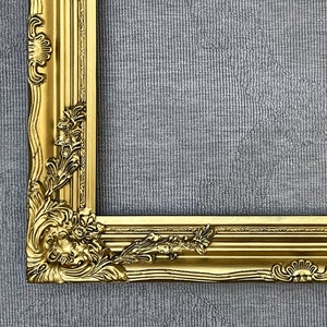 May include: A gold-colored ornate picture frame with a detailed, carved design. The frame is on a gray textured background.