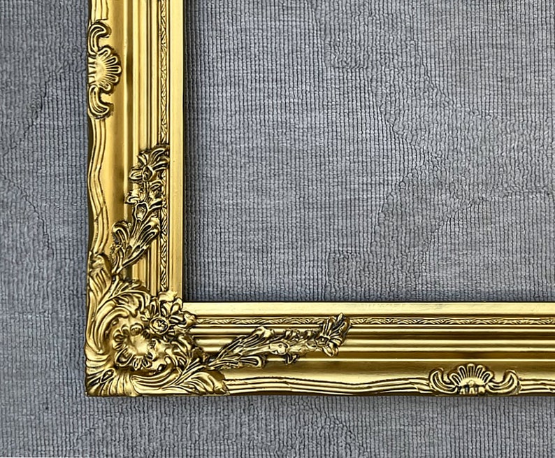 May include: A gold-colored ornate picture frame with a detailed, carved design. The frame is on a gray textured background.