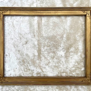 Peut inclure: Un cadre photo rectangulaire doré et orné. Le cadre présente un design détaillé et superposé avec des accents décoratifs aux coins. Le cadre est vide, avec un fond blanc texturé visible.