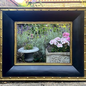 Könnte beinhalten: Ein schwarzer Bilderrahmen mit einem goldenen Seil-Design und goldenen Nieten am Rand. Der Rahmen steht auf einer Betonfläche vor einem Garten.
